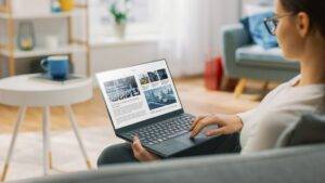 Junge Frau zu Hause verwendet Laptop-Computer zum Scrollen und Lesen von Nachrichten über Wohnungen in Düsseldorf. Sie sitzt auf einer Couch in seinem gemütlichen Wohnzimmer. Über die Schulter geschossen 