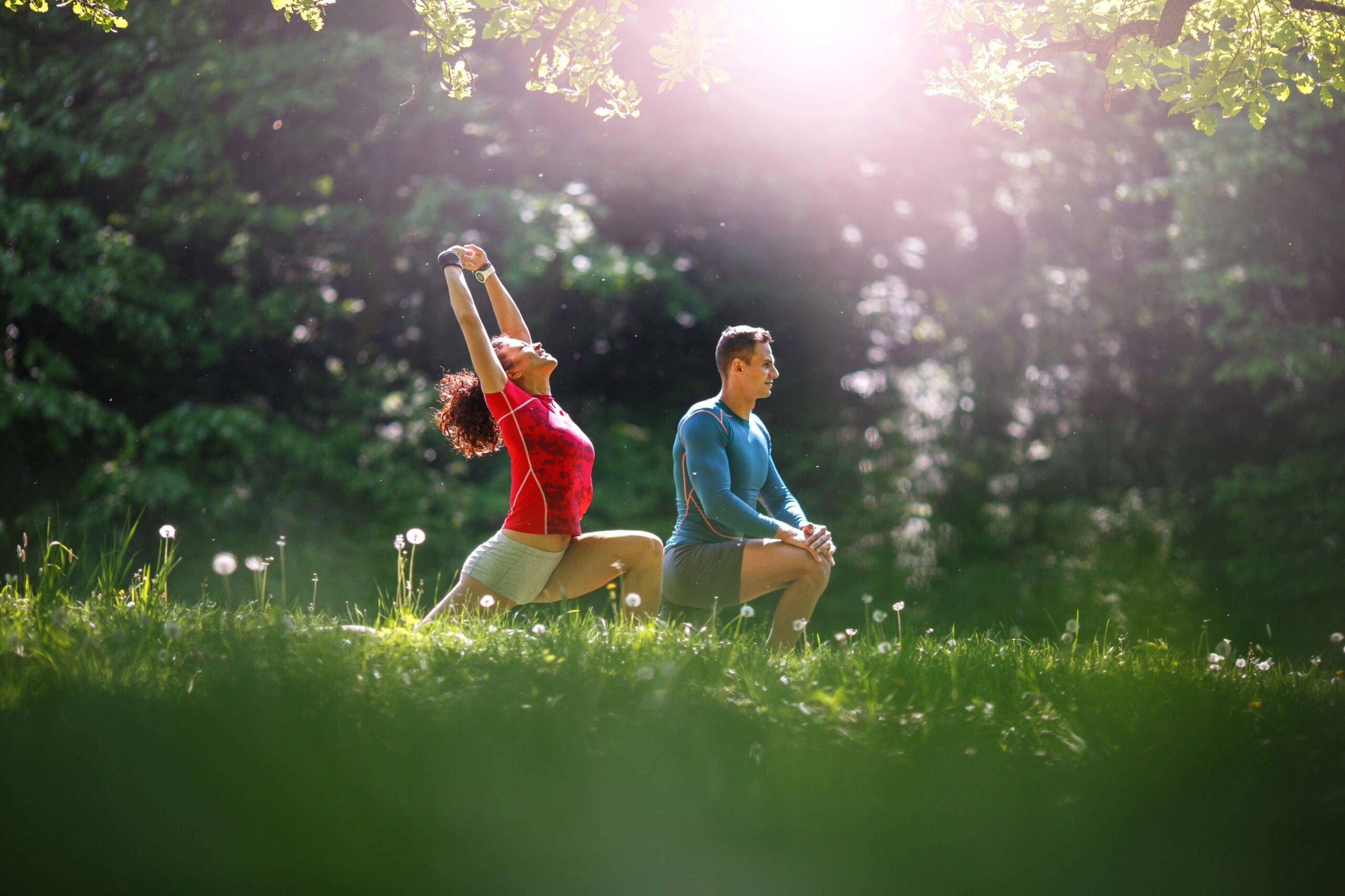Eine Frau und ein Mann machen Dehnübungen in einem grünen Park in Köln, begleitet von einem Personal Trainer in Köln bei Sonnenschein