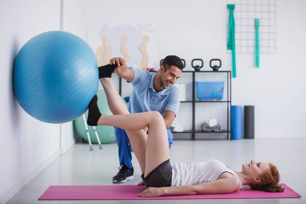 Eine Frau macht Übungen mit einem Gymnastikball im Fitnessstudio in Köln, unterstützt von einem lächelnden Personal Trainer