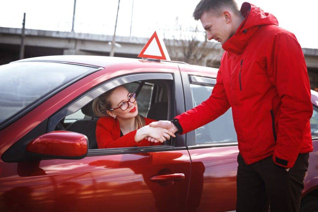 Ein Fahrlehrer in roter Jacke schüttelt die Hand seiner Fahrschülerin, die im Auto sitzt und durch das geöffnete Fenster grüßt. Beide tragen rote Kleidung, während das Fahrzeug mit einem Übungsschild ausgestattet ist. Die Szene symbolisiert den erfolgreichen Abschluss einer Fahrstunde im Rahmen der Fahrlehrerausbildung in der Schweiz