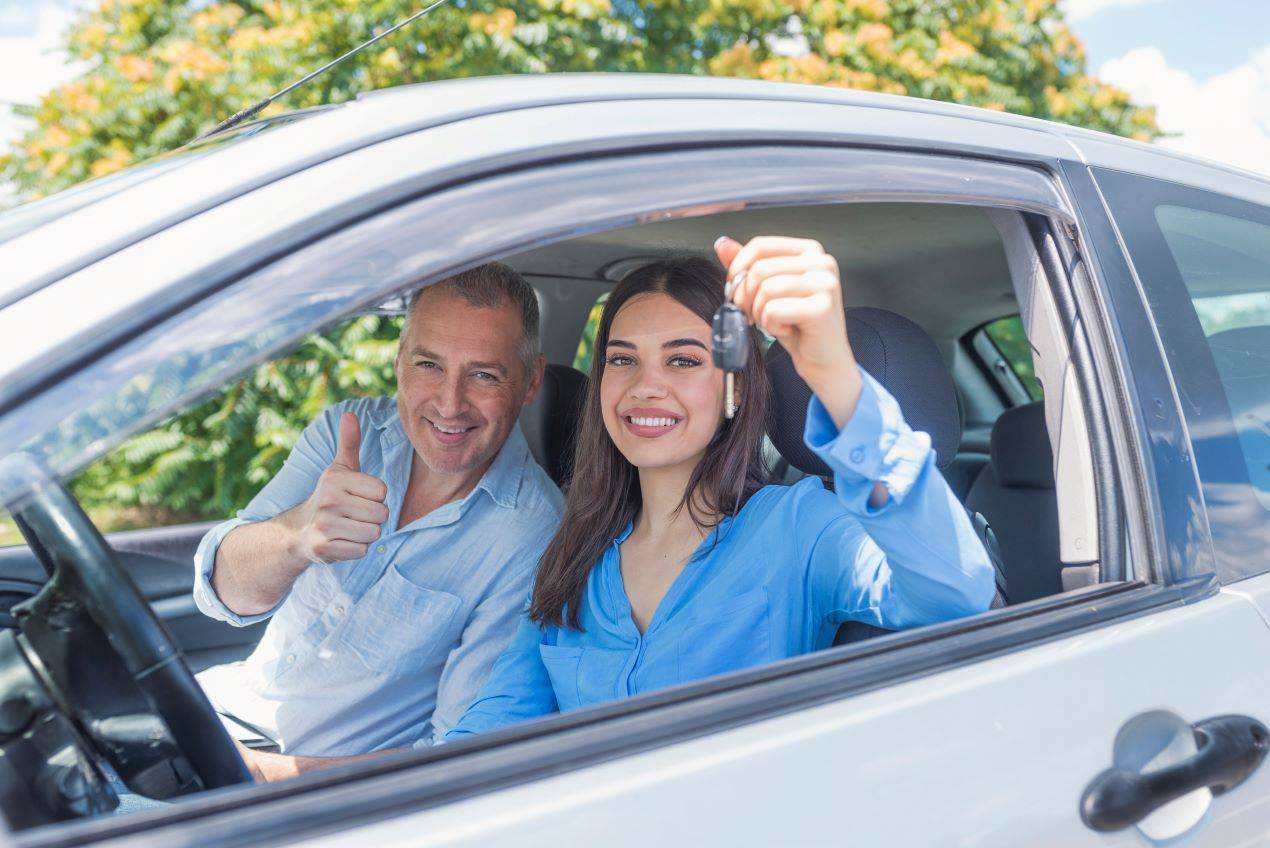 Eine junge Frau hält den Autoschlüssel stolz in der Hand, während ihr Fahrlehrer im Auto neben ihr sitzt und den Daumen nach oben zeigt. Die Szene symbolisiert den Erfolg nach einer Fahrstunde im Rahmen eines Fahrlehrer-Weiterbildungskurses in der Schweiz