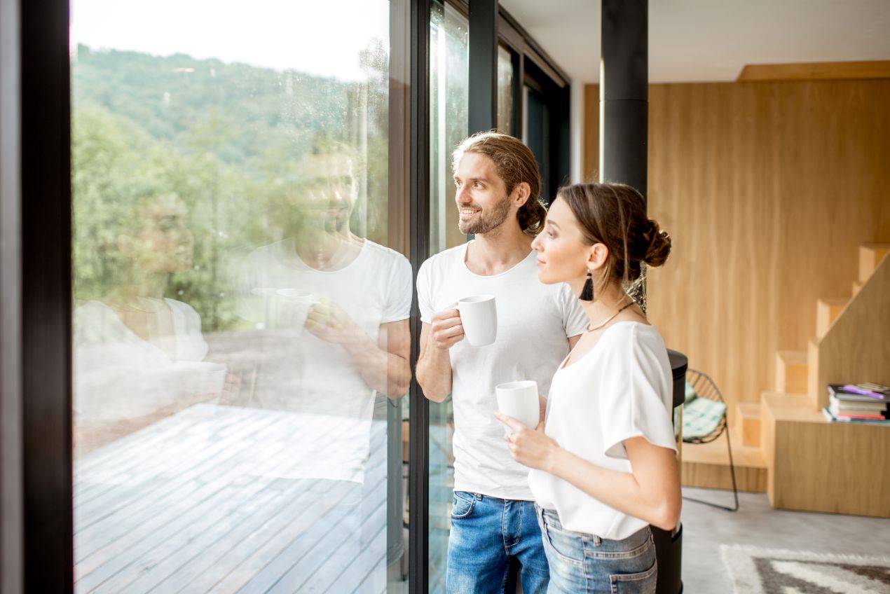 Ein glückliches Paar mit Kaffeetassen steht entspannt vor einer großen Fensterfront mit Fensterfolie blickdicht, während Tageslicht den Raum durchflutet.