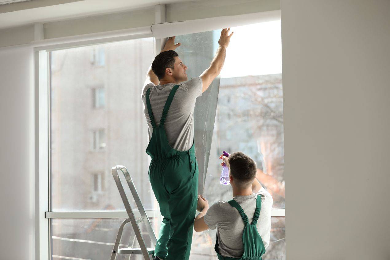 Zwei Handwerker in grünen Overalls montieren eine Fensterfolie blickdicht an einer großen Glasfront in einem modernen Gebäude.